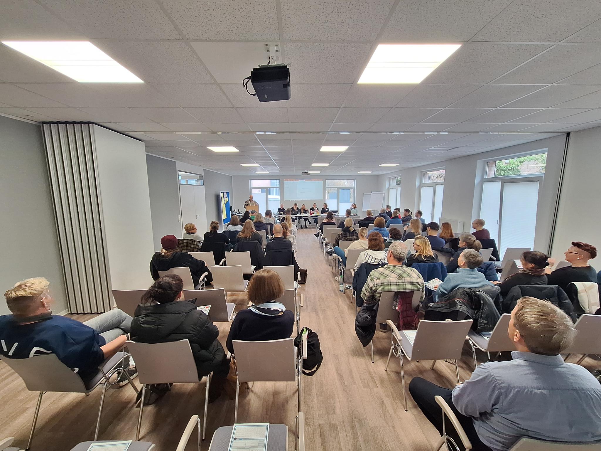 Mehrere Mitarbeitende der Jugendhilfe Collstede sitzen in einem Veranstaltungsraum auf Stuhlreihen und verfolgen aufmerksam eine Präsentation während der Mitarbeitendenversammlung.